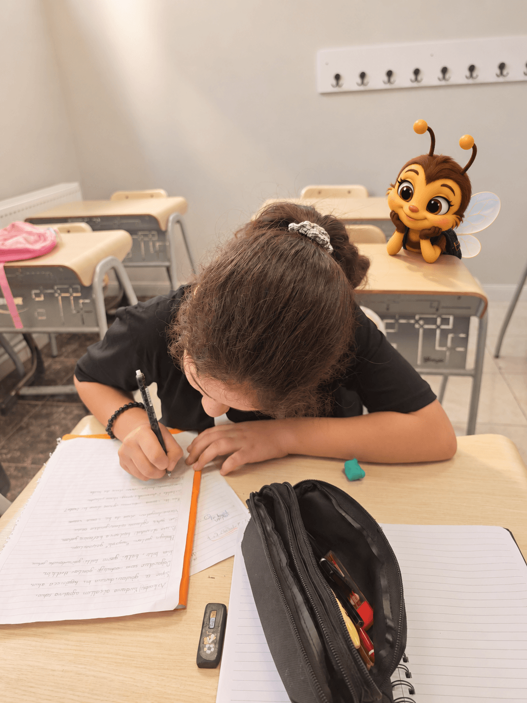 Siyah üst giyen bir çocuk, başını eğerek sıradaki deftere kalemle yazıyor; önünde siyah kalemlik, arka sırada arı maskotu var.
